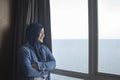 Happy young Asian muslim woman smiling while facing large glass window looking at peaceful ocean view Royalty Free Stock Photo