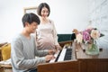 Happy young asian pregnant woman stand side of man play piano at home. Pregnancy, motherhood, people, love, expectation, family - Royalty Free Stock Photo