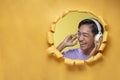 Happy young Asian teenager man laughing poses through torn yellow paper hole with a copy space, wearing purple t-shirt and Royalty Free Stock Photo