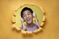 Happy young Asian teenager man laughing poses through torn yellow paper hole with a copy space, wearing purple t-shirt and Royalty Free Stock Photo