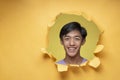 Happy Young Asian teenager man smiling poses through torn yellow paper hole, wearing purple t-shirt with a copy space Royalty Free Stock Photo