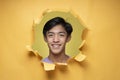 Happy Young Asian teenager man smiling poses through torn yellow paper hole, wearing purple t-shirt with a copy space Royalty Free Stock Photo