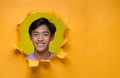 Happy Young Asian teenager man smiling poses through torn yellow paper hole, wearing purple t-shirt with a copy space Royalty Free Stock Photo