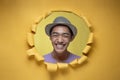 Happy Young Asian teenager man smiling poses through torn yellow paper hole, wearing purple t-shirt and hat Royalty Free Stock Photo