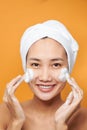 Happy young Asian woman applying face cream while wearing a towel and touching her face. Isolated on orange background Royalty Free Stock Photo