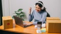 Happy young asian woman checking sales on smartphone and working on laptop computer from home on table with parcel, e-commerce Royalty Free Stock Photo