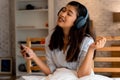 Happy young Asian woman in earphones singing and listening to music while sitting on bed and holding mobile phone at Royalty Free Stock Photo