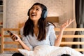 Happy young Asian woman in earphones singing and listening to music while sitting on bed and holding mobile phone at Royalty Free Stock Photo