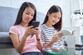 Happy young Asian woman with friends sitting on sofa and reading book, using smartphone in her living room, vacation time. Royalty Free Stock Photo