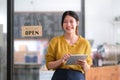 Happy young Asian woman owner standing holding a tablet with open sign board. Royalty Free Stock Photo