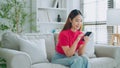 Happy young asian woman relax on comfortable couch at home texting messaging on smartphone