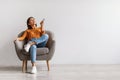 Happy young Asian woman with remote relaxing under air conditioner, sitting in armchair against white wall, mockup Royalty Free Stock Photo