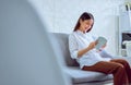 Happy young Asian woman sitting on sofa and reading book in her living room, vacation time. Royalty Free Stock Photo