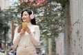 Happy young Asian woman smiling looking at mobile phone wearing headphones and long sleeves sweater while walking down the street Royalty Free Stock Photo