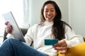 Happy young Asian woman using digital tablet and credit card to shop online sitting on the sofa at home. Copy space. Royalty Free Stock Photo