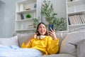 Happy young asian woman wearing headset and relaxing on comfortable couch at home, texting messaging on smartphone