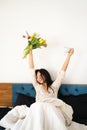 Happy young Asian woman wearing pajamas holding cup and tulips flowers on the bed having fun Royalty Free Stock Photo
