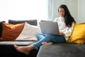 Happy young Asian woman working with laptop at home sitting on the couch in cozy apartment living room. Copy space. Royalty Free Stock Photo