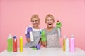 Happy young attractive blonde cleaning ladies dressed in uniform raising hands with spray bottles and smiling gladly at camera,