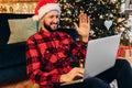 Happy young man, wearing a Santa Claus hat, uses a laptop, talks by video call, enjoys Christmas at home against the background of
