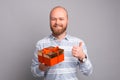 Happy young bearded man holding red gift box and showing thumb up over grey background Royalty Free Stock Photo