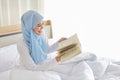 Happy young beautiful asian arab woman in sleepwear sitting, reading book on bed. Cheerful and cute muslim girl resting in bedroom Royalty Free Stock Photo