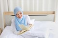 Happy young beautiful asian muslim woman in sleepwear sitting, reading book on bed. Cheerful and cute girl resting in bedroom Royalty Free Stock Photo