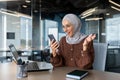 Happy young beautiful muslim woman in hijab working in office at desktop and looking enthusiastically at phone screen Royalty Free Stock Photo