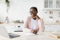 Happy young black lady in glasses and casual sits at table looks at laptop makes notes on minimalist white kitchen