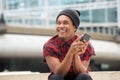 Happy young black man with beanie sitting outside in city with cellphone and looking away Royalty Free Stock Photo