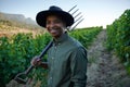 Happy young black man in casual clothing holding pitchfork next to crops on farm