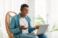 Happy young black man in casual with headphones looks at laptop, has video call, sits in chair Royalty Free Stock Photo