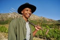 Happy young black man in shirt and brimmed hat holding pitchfork next to plants on farm