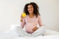 Happy young black pregnant woman holding apple while sitting on white bed Royalty Free Stock Photo