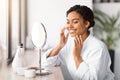 Happy young black woman applying facial moisturizer while looking in makeup mirror Royalty Free Stock Photo