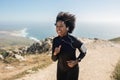 Happy young black woman running in morning, training on rocks with ocean view, enjoying workout outdoors Royalty Free Stock Photo