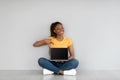 Happy young black woman sitting on floor, pointing at laptop with empty screen against grey wall, mockup for website Royalty Free Stock Photo