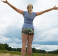 Happy young blond woman, dressed in shorts and a t-shirt stands on the road against the sky arms outstretched Royalty Free Stock Photo