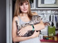 Happy young blonde woman in casual t-shirt holding gorgeous scottish fold cat at domestic kitchen. Natural window light portrait. Royalty Free Stock Photo