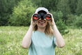 Happy Young blonde woman bird watcher in cap and blue t-shirt looking through binoculars at cloudy sky in summer forest Royalty Free Stock Photo