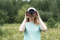 Happy Young blonde woman bird watcher in cap and blue t-shirt looking through binoculars at cloudy sky in summer forest Royalty Free Stock Photo