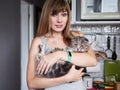 Happy young blonde woman in casual t-shirt holding gorgeous scottish fold cat at domestic kitchen. Natural window light portrait. Royalty Free Stock Photo