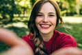 Happy young blonde woman smiling broadly, wearing red sweater, and yellow hat, making self portrait on her device against nature Royalty Free Stock Photo