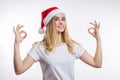 Happy young blonde woman wears red Santa hat and shows OK sign with two hands on white background with copy space