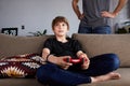 Happy young boy playing video game console seated on a sofa while his father standing close in living room at home Royalty Free Stock Photo