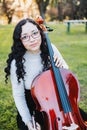 Happy young brunette woman with glasses smiling and playing cello at sunset in the park. Vertical. Royalty Free Stock Photo