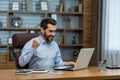 Happy young business man sitting in the office at the desk with a notebook and rejoices at the success, looking at the Royalty Free Stock Photo