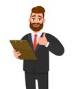 Happy young business man wearing a suit holding clipboard and making or showing thumbs up gesture or sign. Person keeping the file Royalty Free Stock Photo