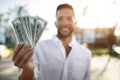 Happy young businessman holding fan of dollar money and smiling at camera, standing outdoors, selective focus, closeup Royalty Free Stock Photo