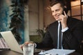 Happy young businessman sitting in cafe using laptop computer talking by mobile phone Royalty Free Stock Photo